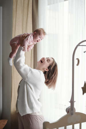 Cheerful young mother playing with her cute baby daughter by cradle with toys