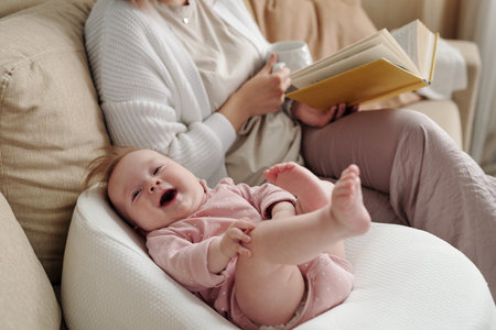 Cute happy infant lying in small bed by her mother with book and mugの写真素材