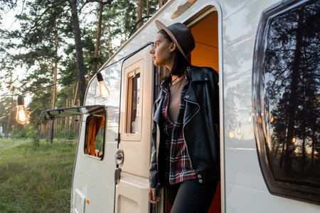Young female traveler in hat and black leather jacket standing in open door of house on wheels in...