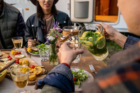 Young man pouring fresh homemade lemonade into glass by served table during outdoor dinnerの写真素材