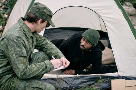 Soldier in army uniform crouching at tent of black man while performing count of migrantsの写真素材