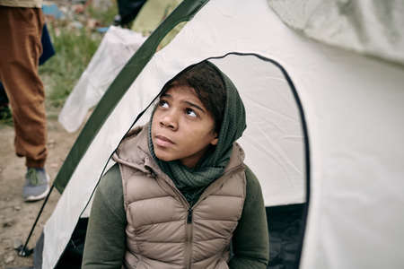 Serious homeless refugee middle-eastern girl in headscarf and vest sitting inside of tent and looking up hopefullyの写真素材