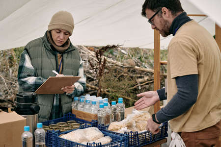 Female volunteer making notes in clipboard while distributing food with assistant to refugeesの写真素材
