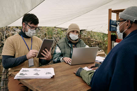 Social workers in masks sitting at table and using devices while talking to black migrant under tentの写真素材