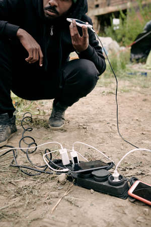 Close-up of black refugee man crouching at socket outlet and listening to audio message on phone while charging it outdoorsの写真素材