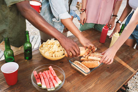 Group of young intercultural friends taking fresh grilled hotdogs while standing by wooden tableの写真素材