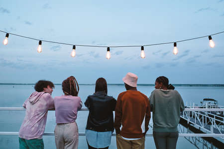 Row of young restful friends standing on pier in front of waterside on summer eveningの写真素材