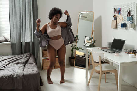 Young cheerful female in underwear and grey shirt dancing in the morning between bed and tableの写真素材