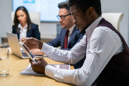 Serious businessman scrolling in tablet while sitting by table against his colleagues