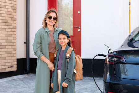 Happy young female and little schoolgirl standing by electric car at charging station