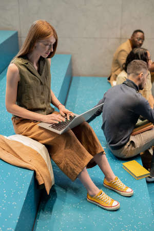 Pretty student typing o laptop keypad while sitting on blue staircase against her groupmatesの写真素材