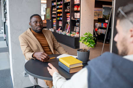 Young African businessman looking at colleague during interaction in bookstore or libraryの写真素材