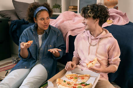 Two intercultural teens discussing something while eating pizza in home environmentの写真素材