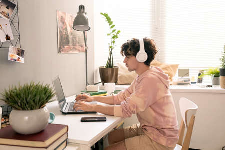 Young man with headphones sitting by desk and networking in front of laptopの写真素材