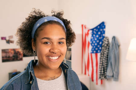 Happy African teenager in casualwear standing on background of American flag in her roomの写真素材