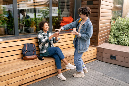 Two happy students relaxing on wooden bench by modern building outdoorsの写真素材