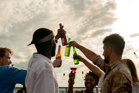 Group of happy friends clinking by bottles of beer against cloudy skyの写真素材
