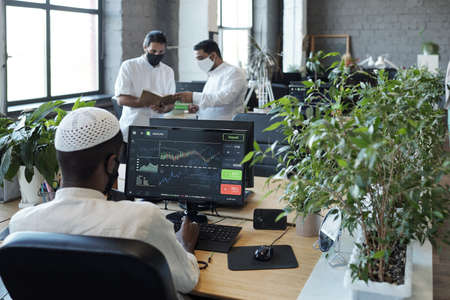 Rear view of young Muslim businessman sitting in front of computer monitor and his two colleagues in officeの写真素材