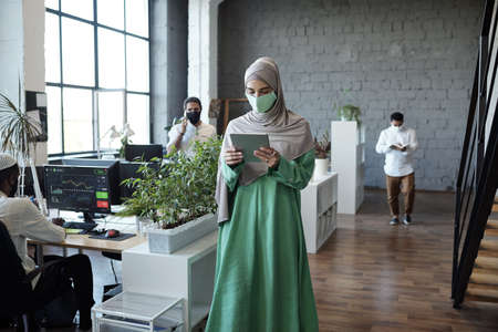 Young businesswoman in hijab looking at tablet screen while walking along tables in large officeの写真素材