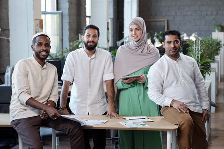 Four young successful intercultural businesspeople standing by workplace in large office