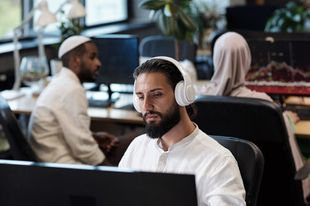 Young man in headphones watching online video against his two colleagues interacting by workplaceの写真素材
