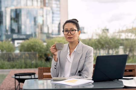 Young businesswoman with cup of coffee sitting in front of laptop by table in outdoor cafeの写真素材