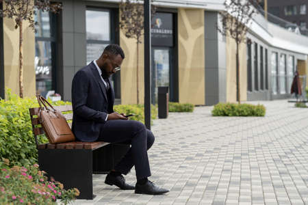 Young bearded African businessman scrolling in smartphone while sitting on benchの写真素材