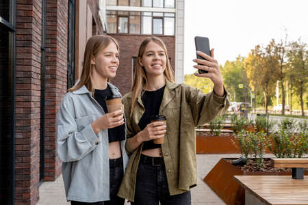 Two cute blond teenage girls with drinks making selfie or communicating through video call outdoors