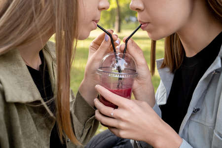 Two thirsty teenage girls sharing fruit cocktail or juice in park on sunny dayの写真素材
