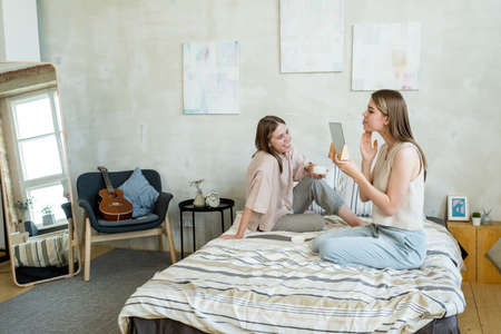 Happy girl with drink looking at her twin sister taking care of her skin while both relaxing on bedの写真素材