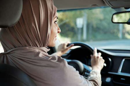Horizontal rear view portrait shot of modern Muslim woman wearing hijab driving car looking into...