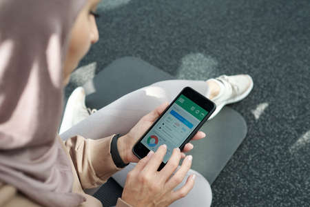 High angle view shot of modern young adult Muslim woman sitting on mat in gym reading information in fitness app on smartphoneの写真素材