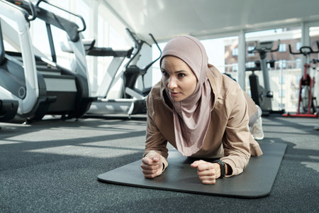 Horizontal medium close-up portrait shot of strong young adult Muslim woman wearing hijab doing elbow plank exercise in gymの写真素材