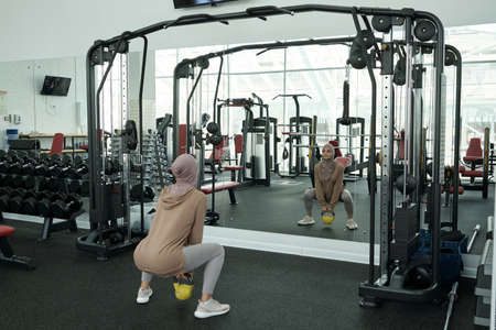 Horizontal long shot of sporty Muslim woman wearing headwrap doing squats exercise with kettlebell...