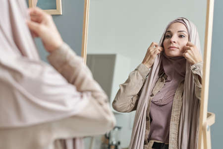 Horizontal over-the-shoulder shot of attractive young adult Muslim woman getting ready to go out putting on hijab in front of mirrorの写真素材