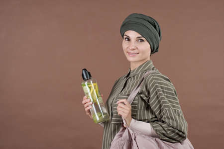 Horizontal medium studio portrait shot of stylish young adult Muslim woman holding bottle of fresh homemade lemonade smiling at camera, brown backgroundの写真素材