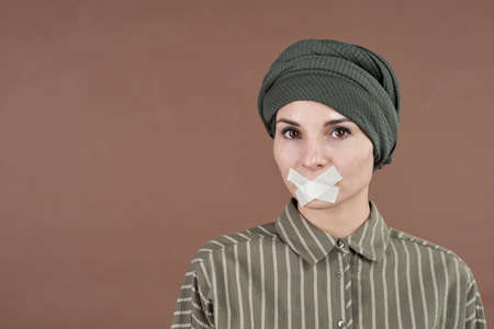 Horizontal medium close-up conceptual portrait shot of Muslim woman wearing turban standing with tape on her mouth looking at cameraの写真素材