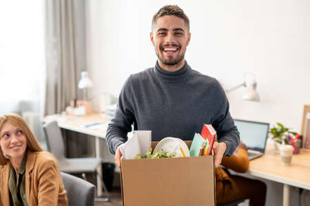 Young cheerful employee with box containing office supplies standing against co-workersの写真素材