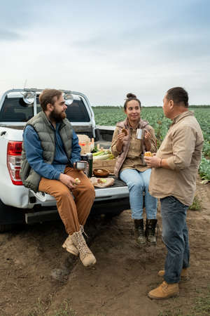 Three contemporary farmers having lunch on car trunk against cabbage fieldの写真素材