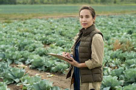 Young confident female farmer making notes in document against cabbage field