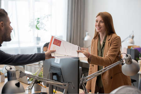 Young smiling businesswoman passing financial papers to male colleague over computer monitorの写真素材