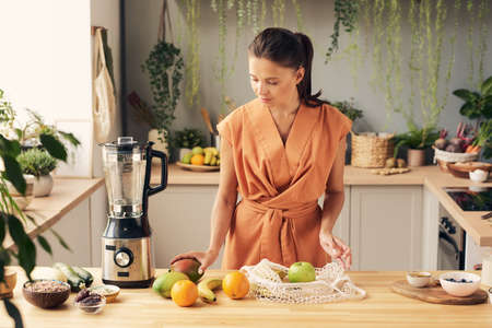 Young house wife preparing fresh fruits while going to make smoothie in electric blenderの写真素材