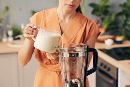 Woman in orange dress pouring fresh milk into electric blender while making smoothieの写真素材
