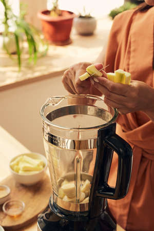 Hands of young female cutting fresh zucchini into electric blender while making smoothie