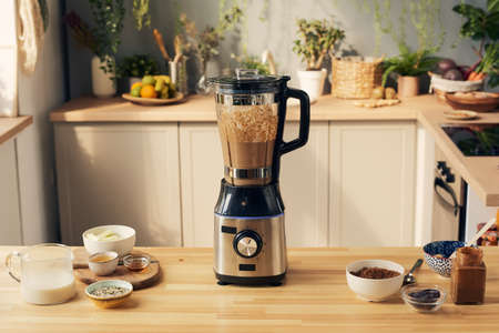 Kitchen table with electric blender with chocolate smoothie and bowls with ingredientsの写真素材