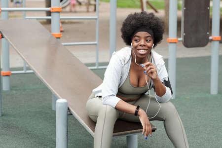 Young smiling sportswoman with earphones communicating through mobile technologies while sitting on sportsgroundの写真素材