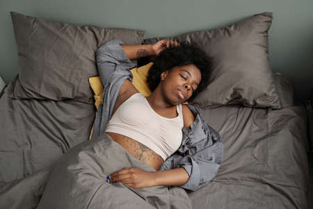Young tired African woman sleeping in double bed with two pillows at nightの写真素材