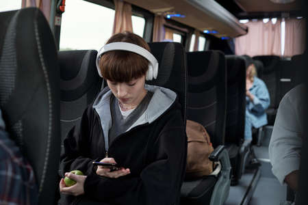 Cute teenage girl in headphones scrolling through playlist in smartphone while sitting in busの写真素材