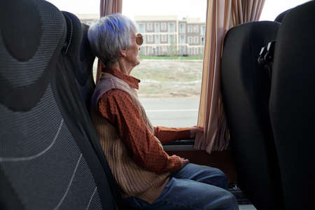 Elderly woman in casualwear looking through window while traveling by intercity busの写真素材
