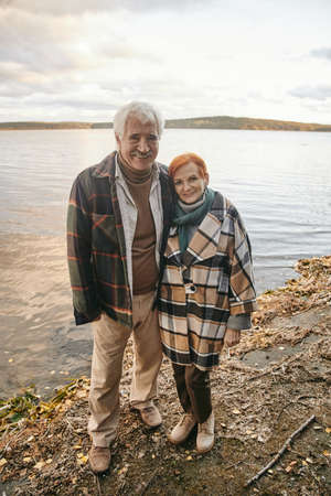 Portrait of happy senior couple embracing and smiling at camera while standing near the lake...
